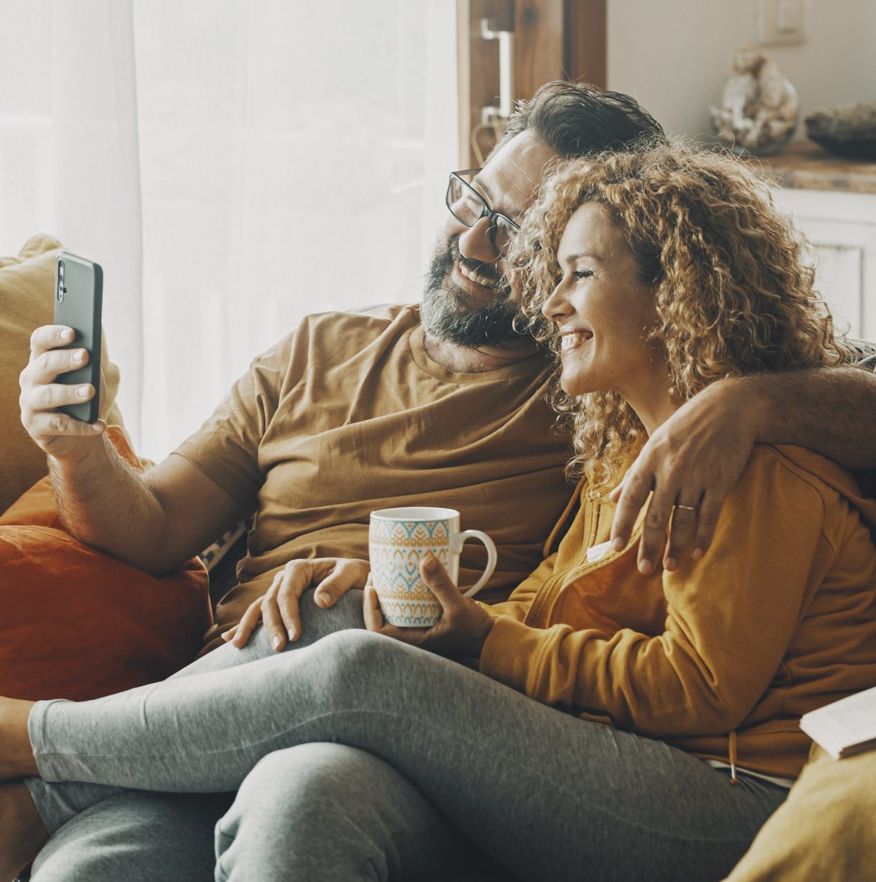 Couple relax on a sofa, smiling as the man holds up a phone to take a selfie.